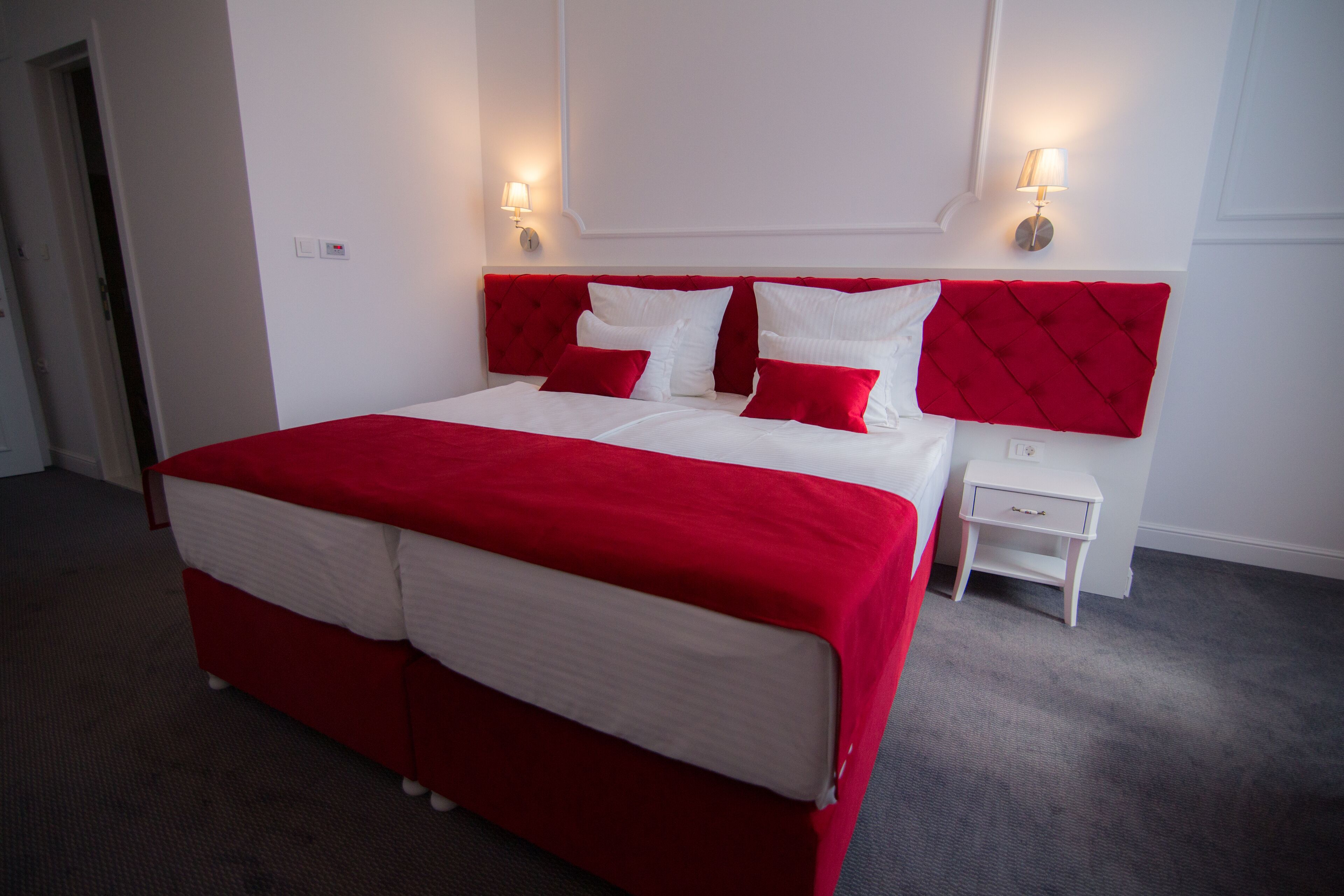Una habitación de hotel con una cama grande, ropa de cama roja y blanca, dos mesitas de noche con lámparas y piso alfombrado.