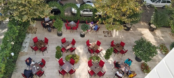 Ein Straßencafé mit mehreren Tischen und Stühlen, umgeben von Kübelpflanzen und Bäumen.