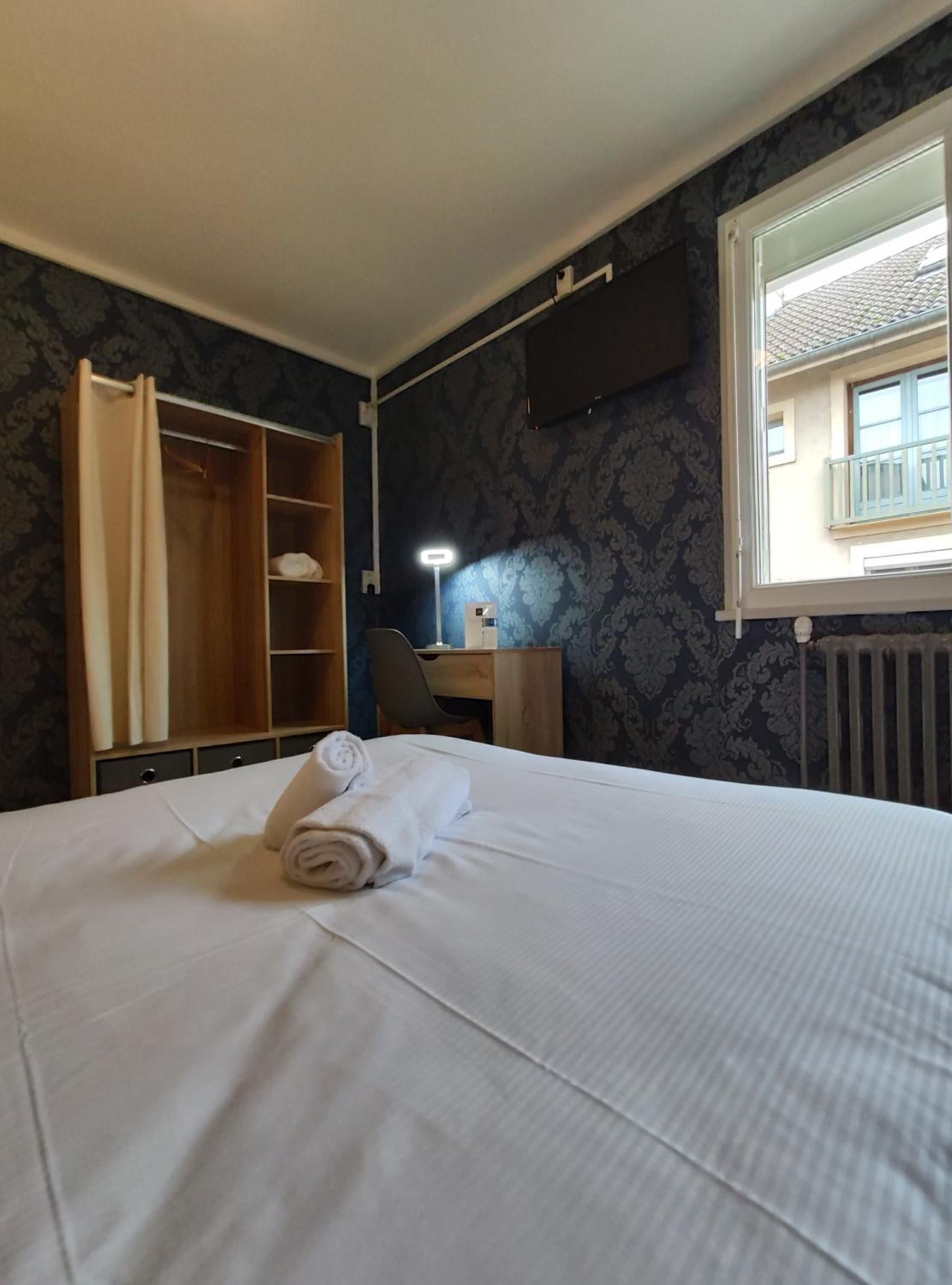 A hotel room with a bed, a desk, a chair, a wardrobe, and a window with a view of a balcony.