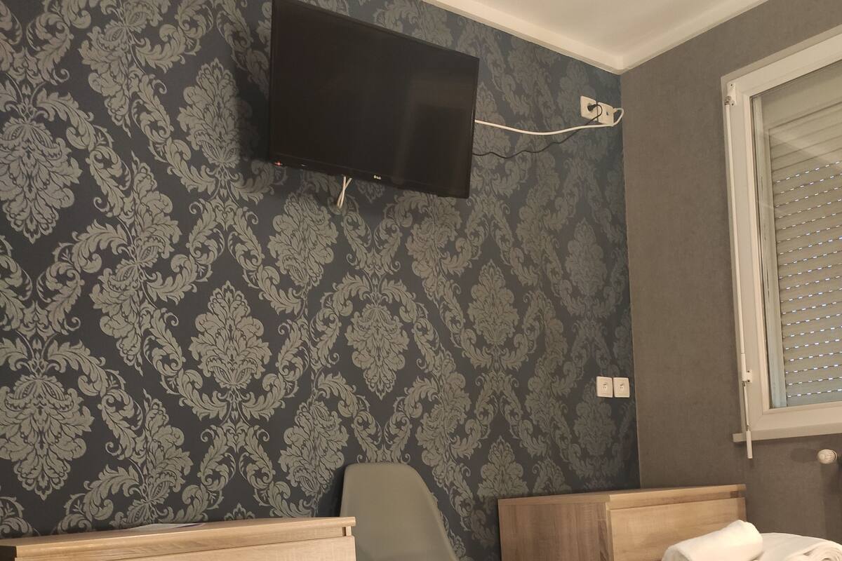 Hotellihuone, jossa on kaksi sänkyä, seinälle asennettu televisio, tuoli ja ikkuna, jossa on sälekaihtimet.