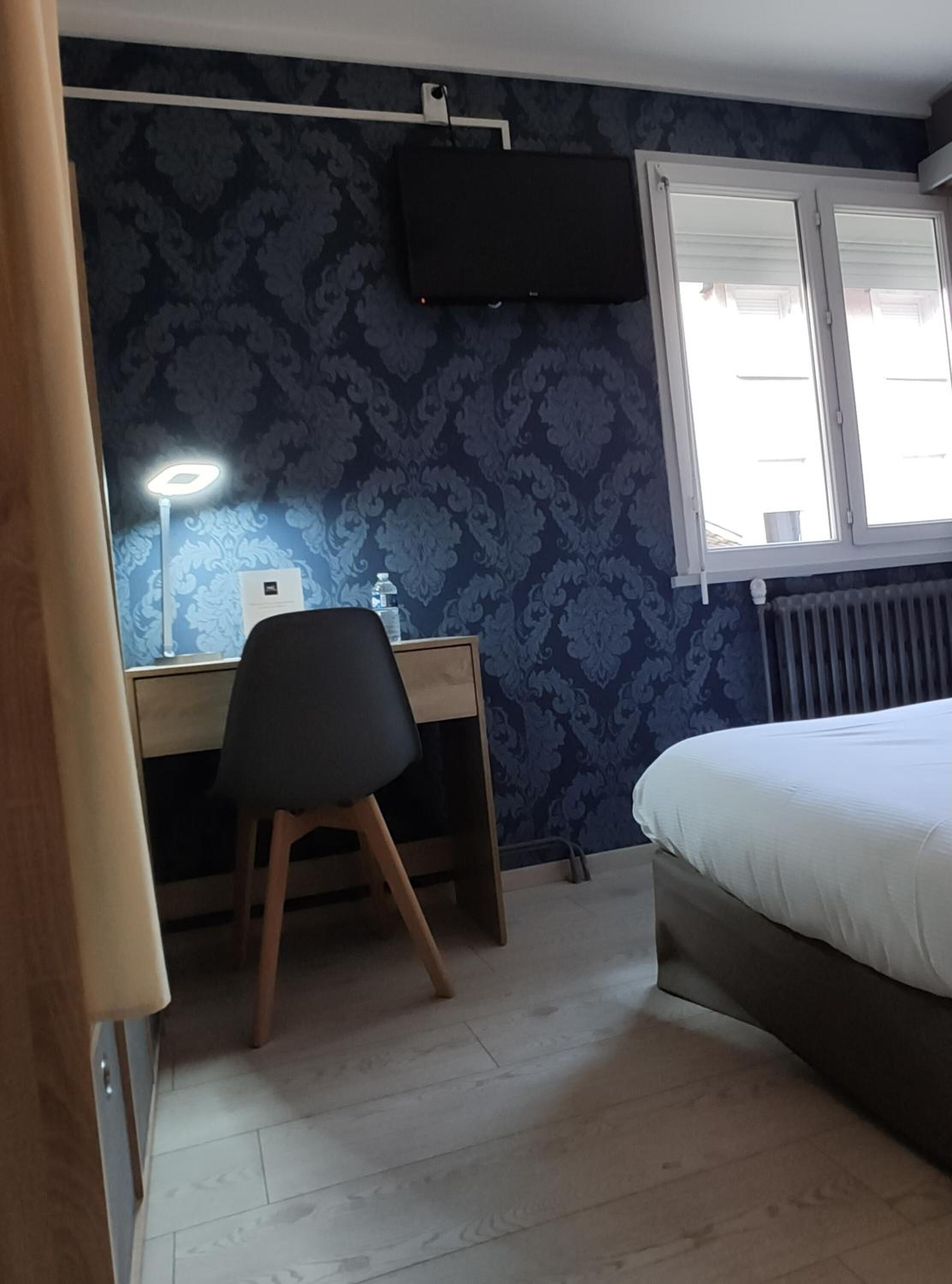Une chambre à coucher comprenant un lit, un bureau avec une chaise, une lampe sur pied et une fenêtre avec des stores.