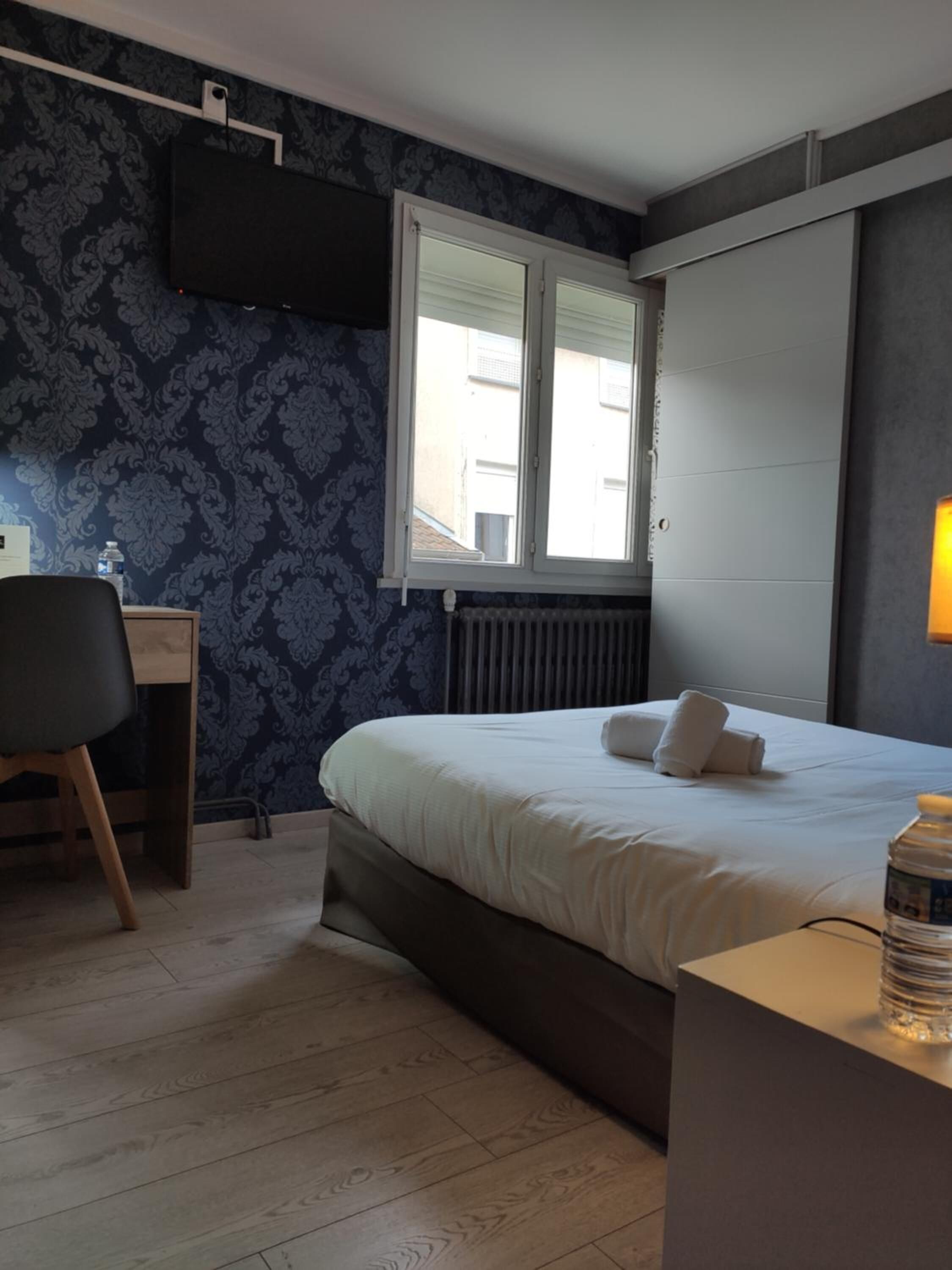 A hotel room with a bed, a desk, a chair, a radiator, and a window with blinds.