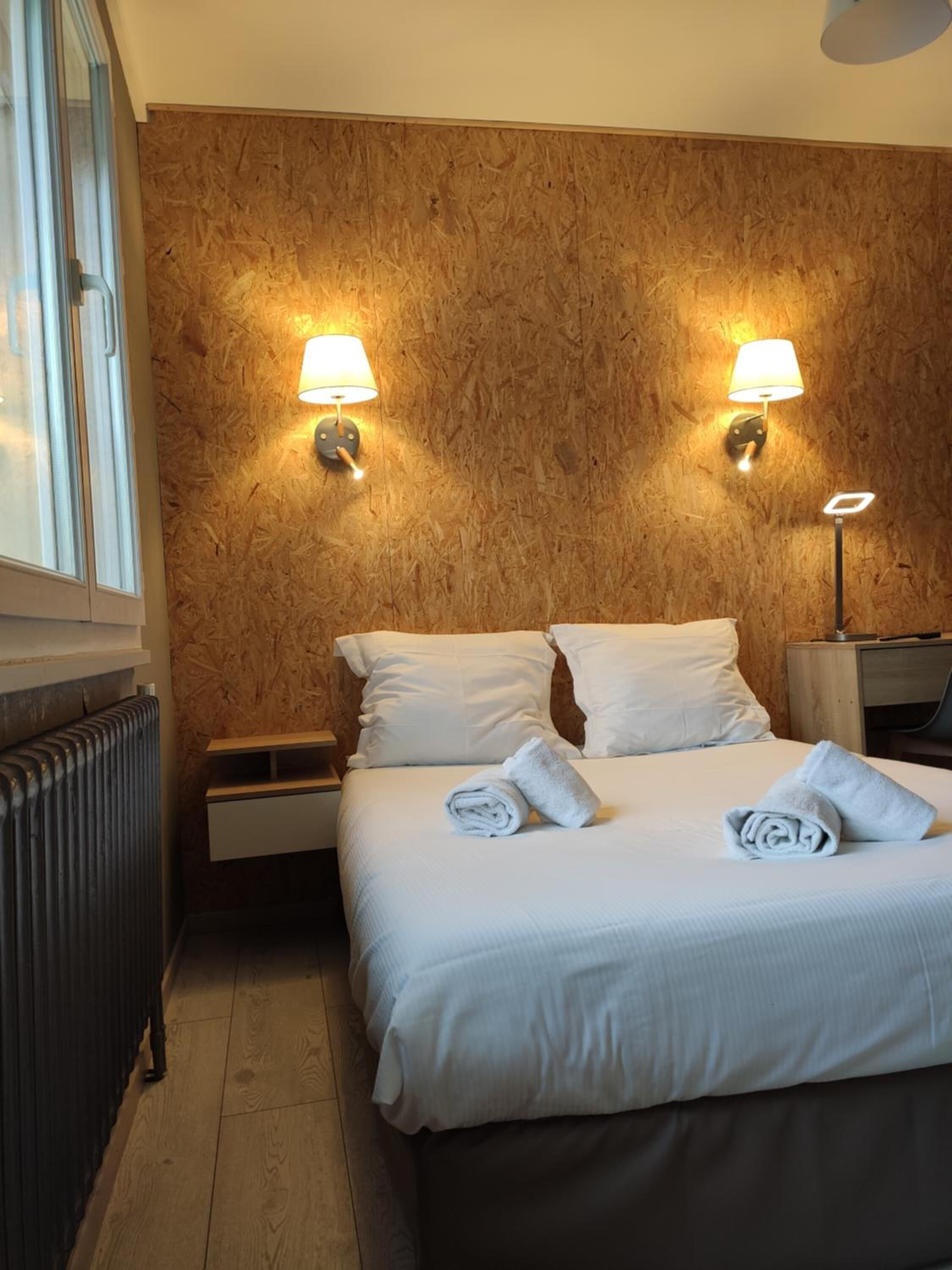 Habitació d'hotel amb una paret de fusta, dues làmpades de nit, un llit amb roba de llit blanca i un radiador.