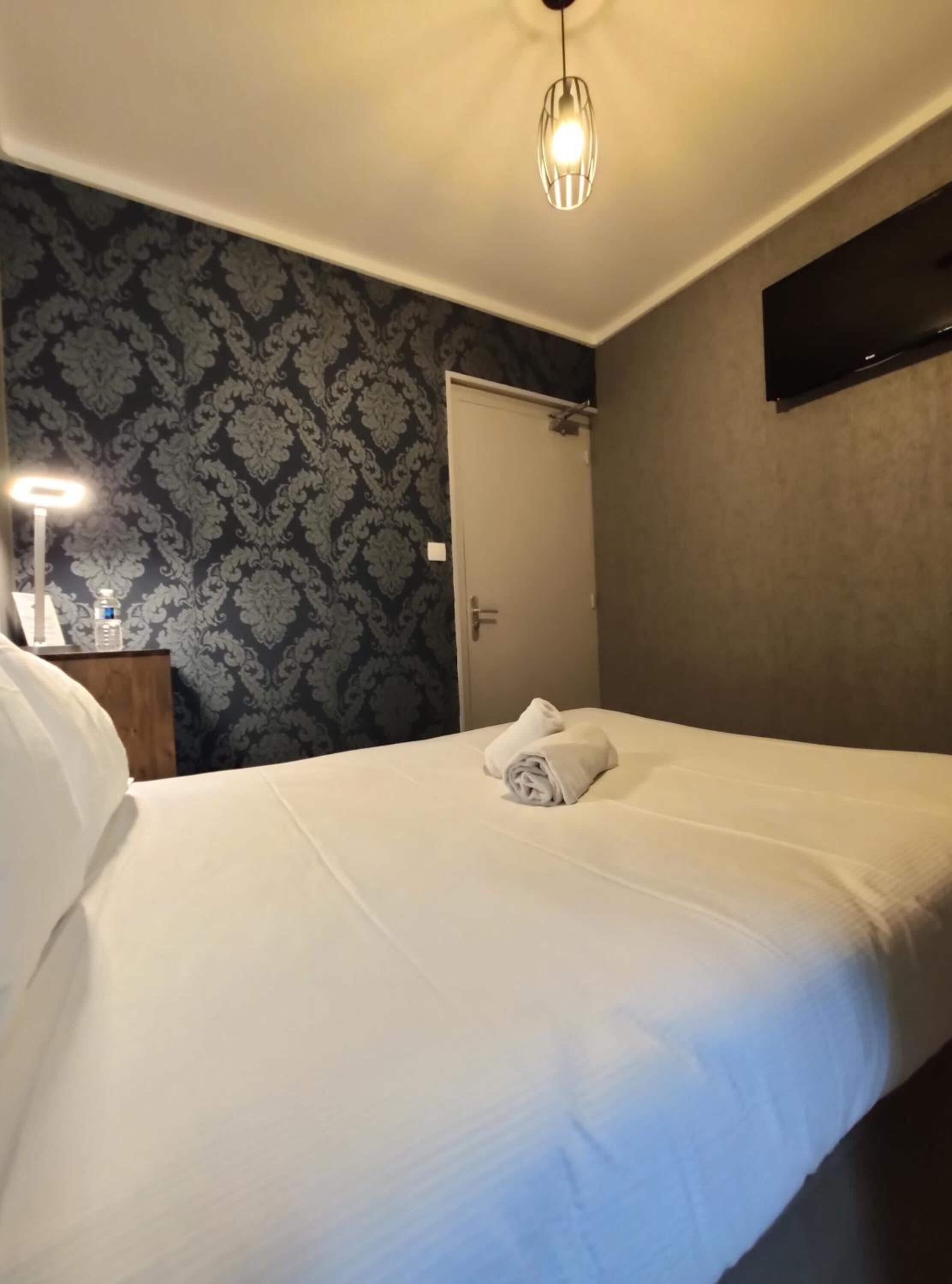 Una habitación de hotel con una cama bien hecha, una mesita de noche con lámpara y un papel tapiz estampado.
