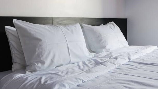 A neatly made bed with white bedding and pillows, a dark headboard, and a plain wall in the background. - Memo's Homestay (Mlolongo)