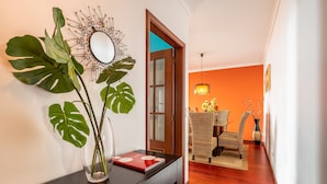 Un pasillo con una consola de madera, un espejo grande y una planta de monstera en maceta. Se ve una puerta que da a una habitación con sillas de mimbre y un florero.
