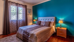 Un dormitorio con una cama grande, una mesita de noche, una lámpara y vistas al exterior a través de una ventana con cortinas.