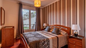 A bedroom with a wooden bed, striped wallpaper, a nightstand with lamps, and a view of the outdoors. - Casa Branca II (Funchal)