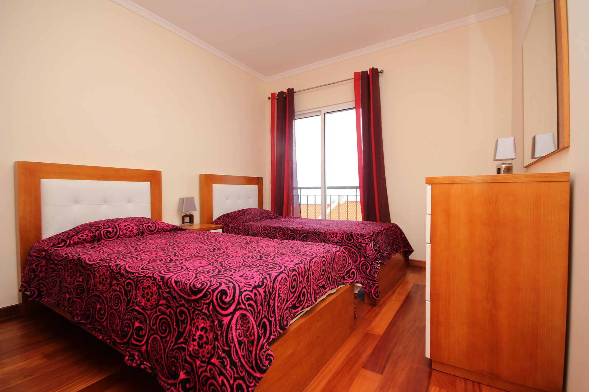 A hotel room with two beds, a wooden headboard, a desk with a lamp, a wardrobe, and a window with curtains.