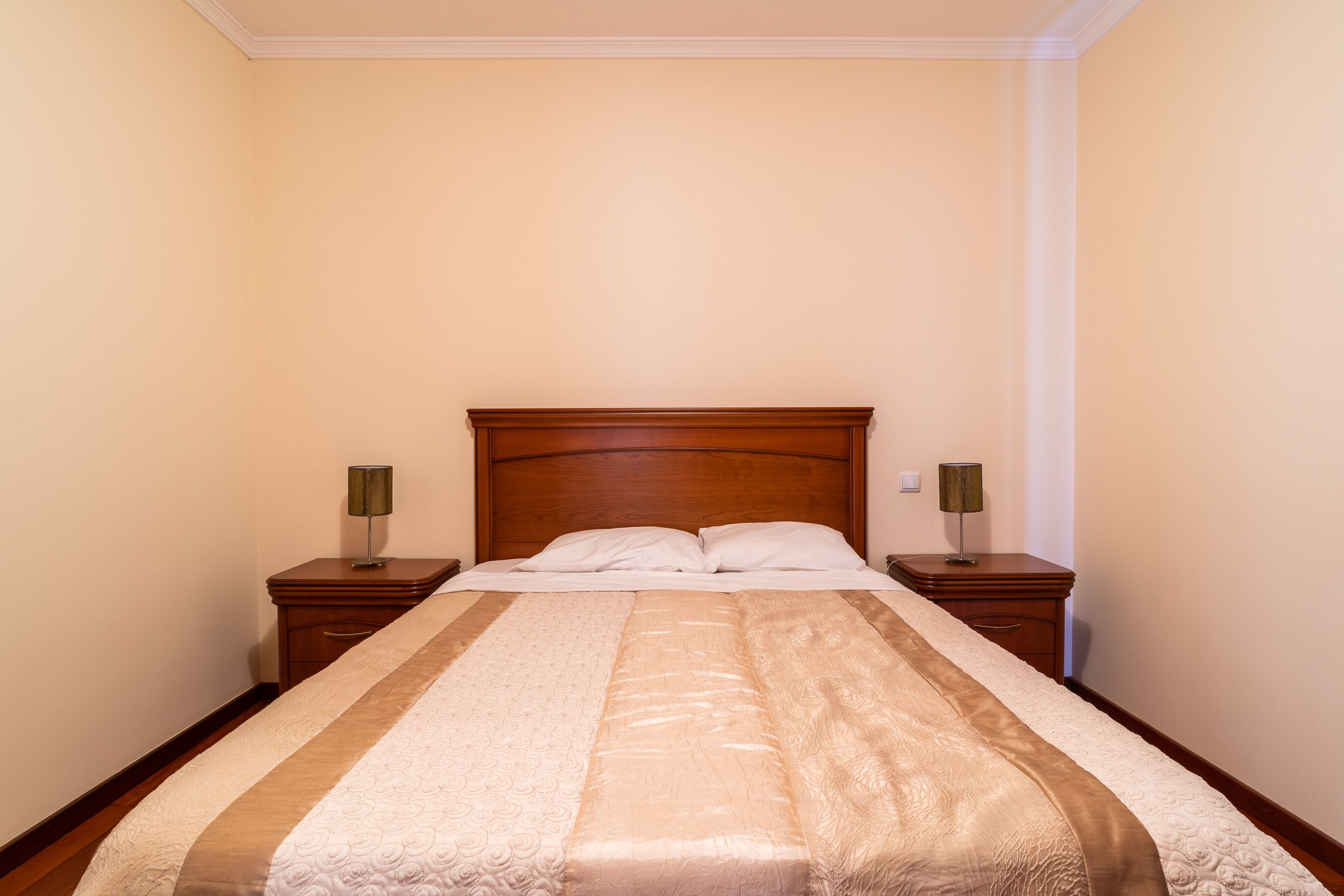 A neatly made bed with a wooden headboard, two bedside tables with lamps, and a plain wall.