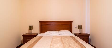 A neatly made bed with a wooden headboard, two bedside tables with lamps, and a plain wall.
