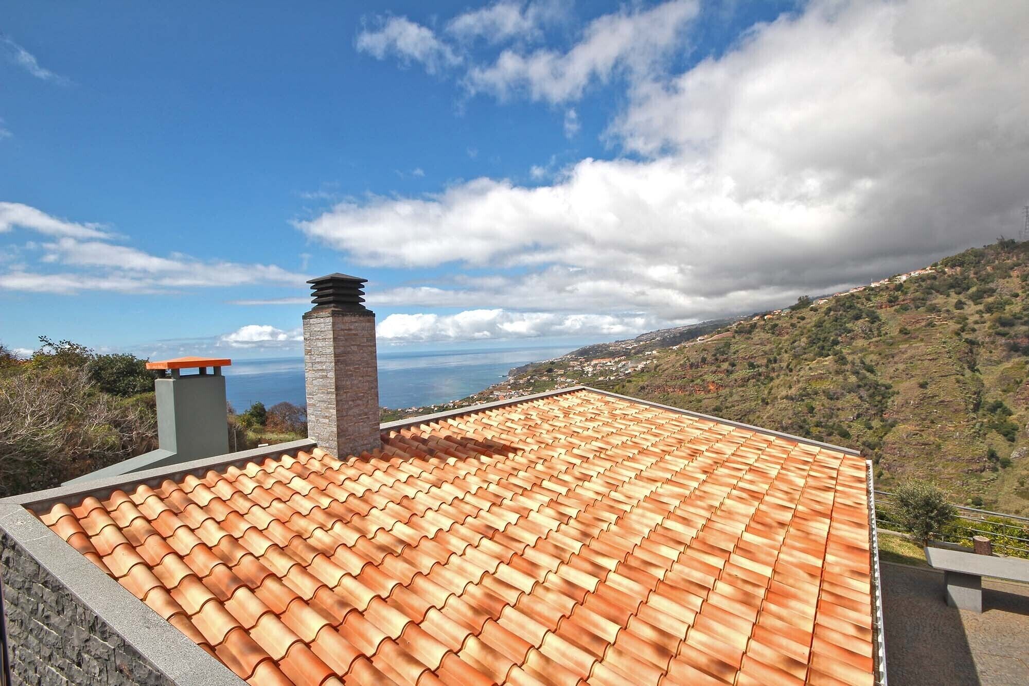 A rooftop with terracotta tiles, a stone chimney, and a view of a coastal landscape.