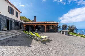 A building with a stone facade, a covered patio with outdoor seating, and a view of the sea. - Gran Horizonte House (Ribeira Brava)