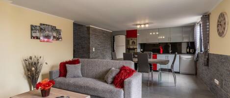 A modern living room with a grey sofa, red throw pillows, and a wooden coffee table. A dining area with a red table and chairs is visible in the background.
