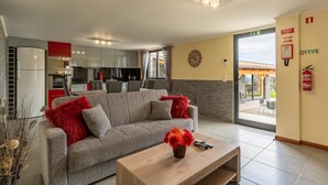 Ein modernes Wohnzimmer mit einem grauen Sofa, roten Zierkissen und einem Holztisch. Im Hintergrund ist eine Küche mit roten Schränken und Edelstahlgeräten zu sehen.