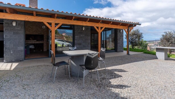 Eine überdachte Terrasse mit Steinboden, einem Tisch mit Stühlen und Blick auf einen Berg sowie ein Auto durch die Glastüren hindurch.