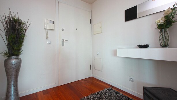 Een moderne kamer met houten vloer, een witte deur, een lichtschakelaar op de muur, een thermostaat, een klein wit plankje met een kom, en een vaas met planten.