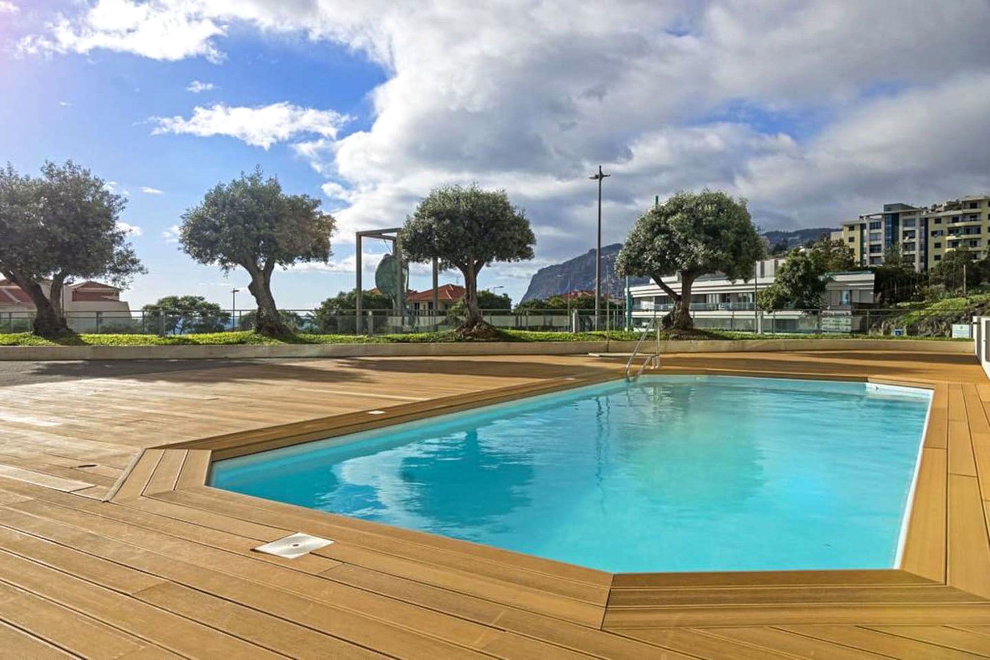 Una piscina rectangular con plataforma de madera, rodeada de árboles y edificios.