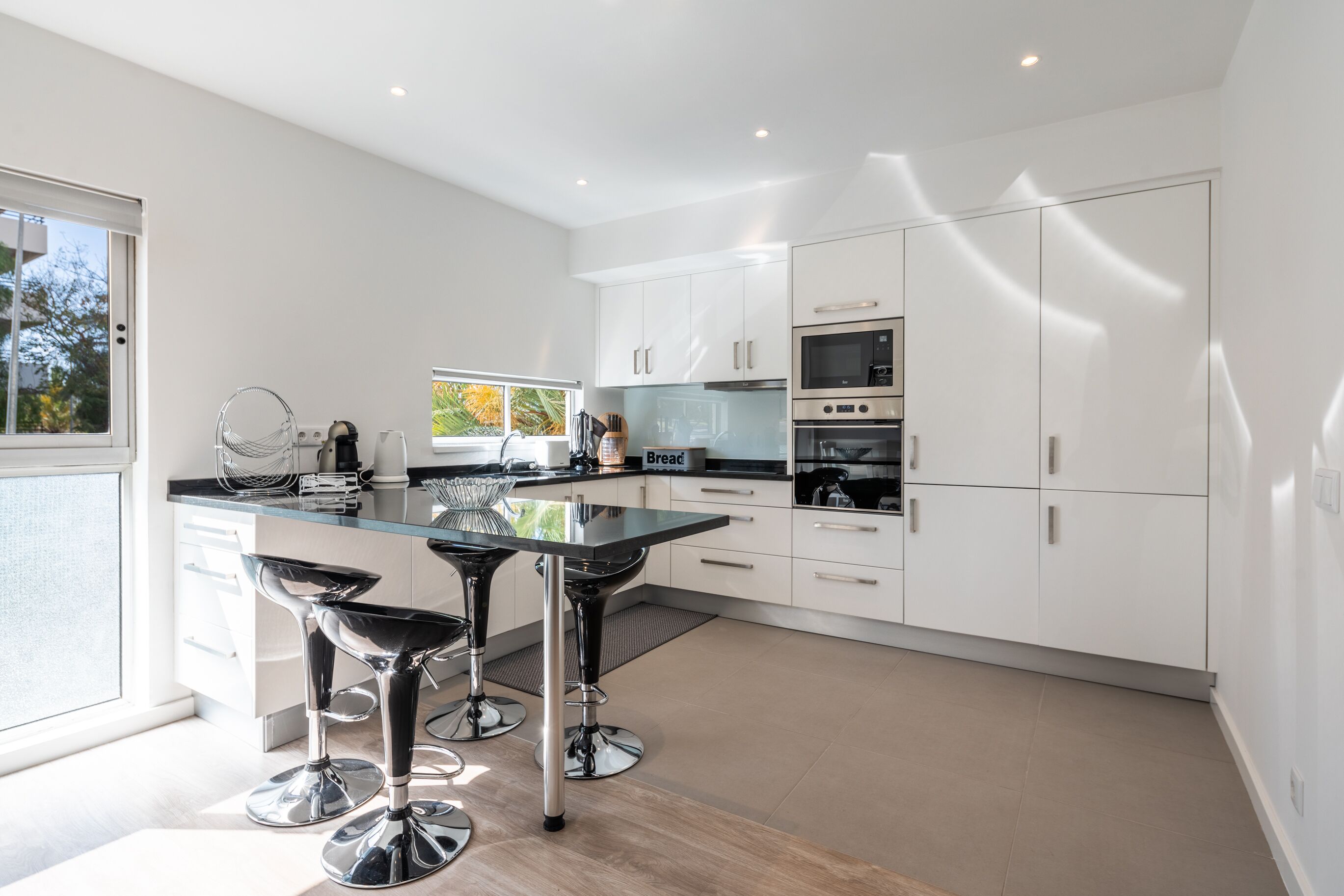 A modern kitchen with white cabinets, a black countertop, and stainless steel appliances. There are two bar stools and a window providing natural light.