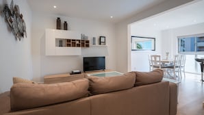 Ein modernes Wohnzimmer mit einem großen beigen Sofa, einem Flachbildfernseher und einem Essbereich mit weißen Stühlen und einem Tisch.