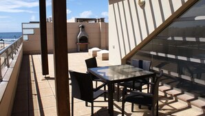 A rooftop terrace with a glass table and chairs, a barbecue grill, and a view of the ocean.