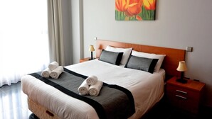 A hotel room with a neatly made bed, a painting of flowers on the wall, and a window with curtains.