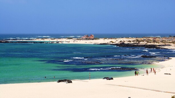 A beach with clear turquoise waters, a sandy shore, and a building with a red roof in the distance.