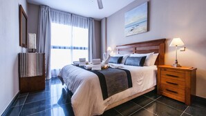 A hotel room with a large bed, a wooden nightstand, a bedside lamp, a painting on the wall, and a window with curtains.