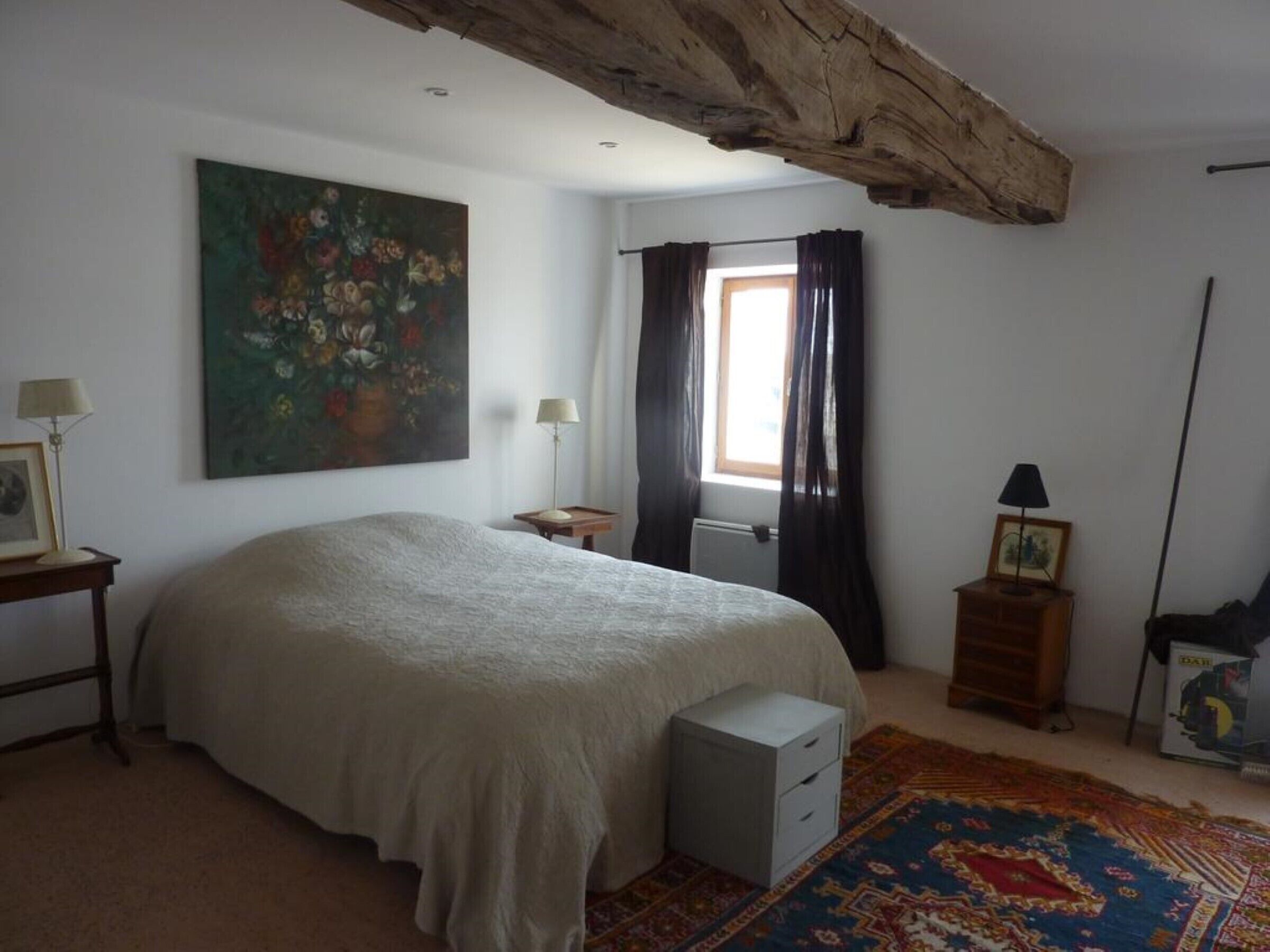 Un dormitorio con una cama grande, un cuadro, una ventana con cortinas, una mesita de noche, una lámpara y una alfombra.