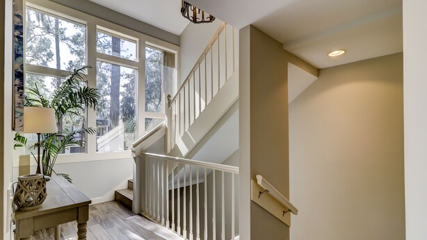 Eine Treppe mit weißen Geländern führt zu einer oberen Etage, ein Holztisch mit Lampe und einer Dekoration, und ein großes Fenster mit Blick auf Bäume.