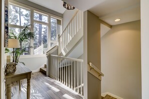 Un escalier avec des rampes blanches menant à l’étage supérieur, une table en bois avec une lampe et un objet décoratif, et une grande fenêtre donnant sur des arbres.