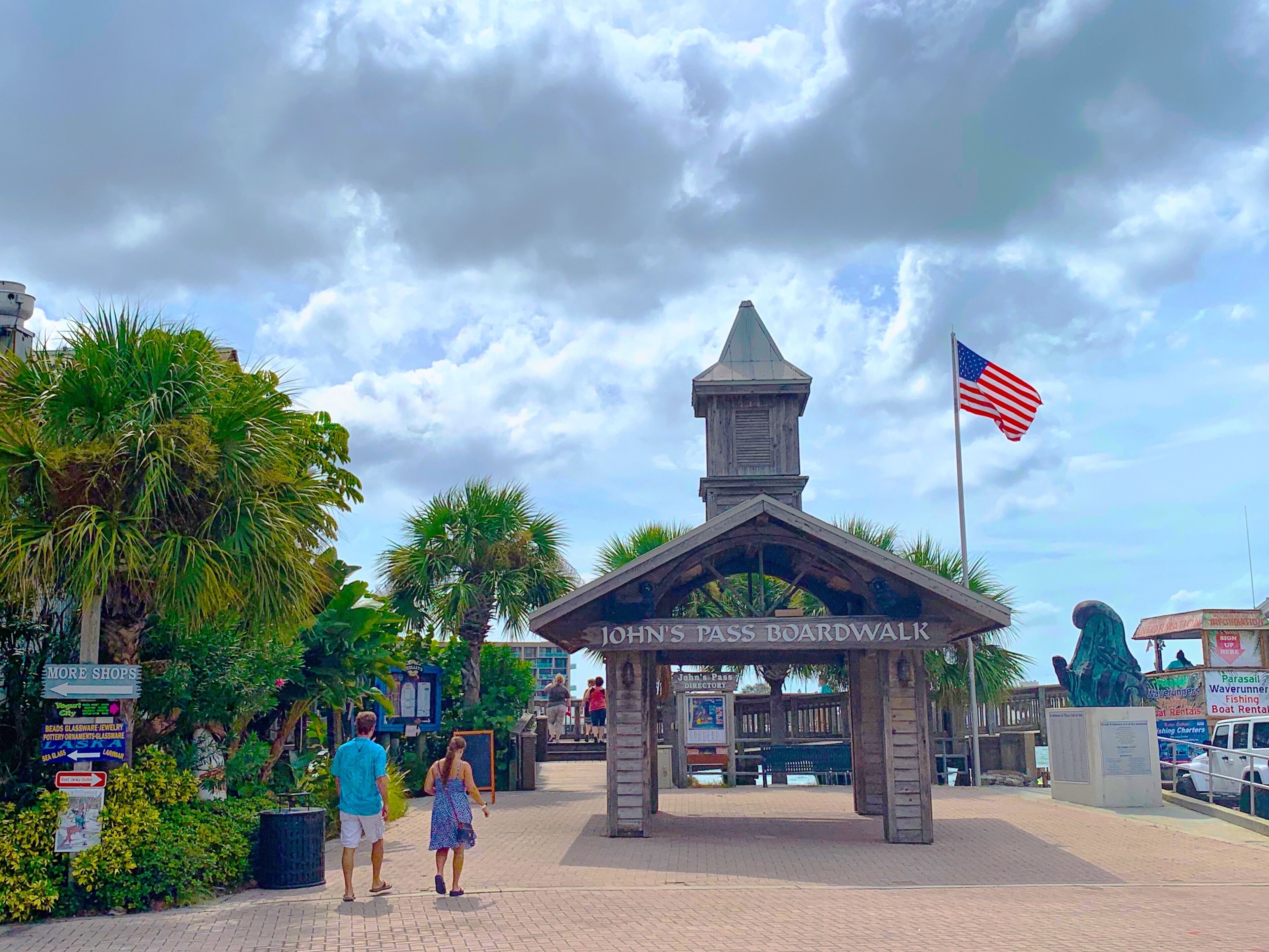 A wooden structure labeled "John's Pass Boardwalk" with an American flag, surrounded by palm trees and a clear sky.