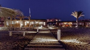 Un hôtel situé en bord de mer, avec un chemin de pierre menant à l’entrée, des palmiers et une structure surmontée d’un toit de chaume.