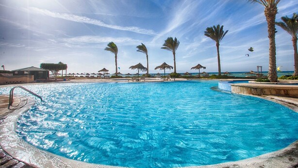Une grande piscine extérieure, avec des palmiers et des parasols de plage en arrière-plan.