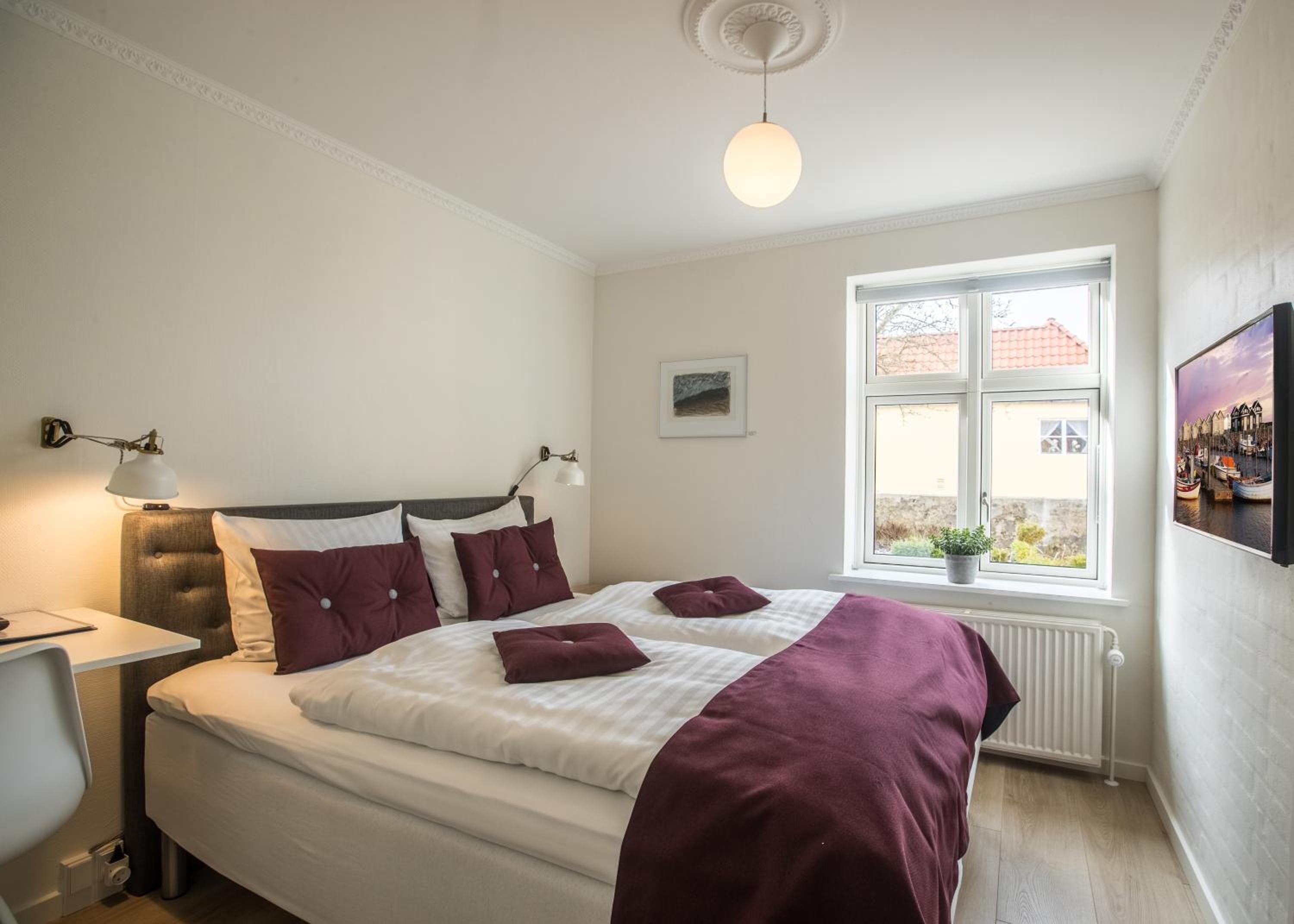 A neatly made bed with a white and maroon bedspread, two bedside lamps, a small potted plant by the window, and a framed picture on the wall.