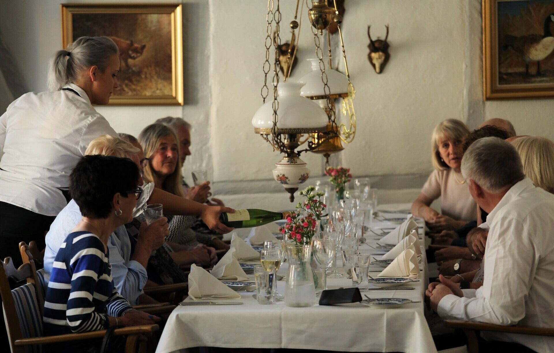 Un elegante ambiente de comedor con invitados sentados alrededor de una mesa, un camarero atendiendo y una araña colgando del techo.