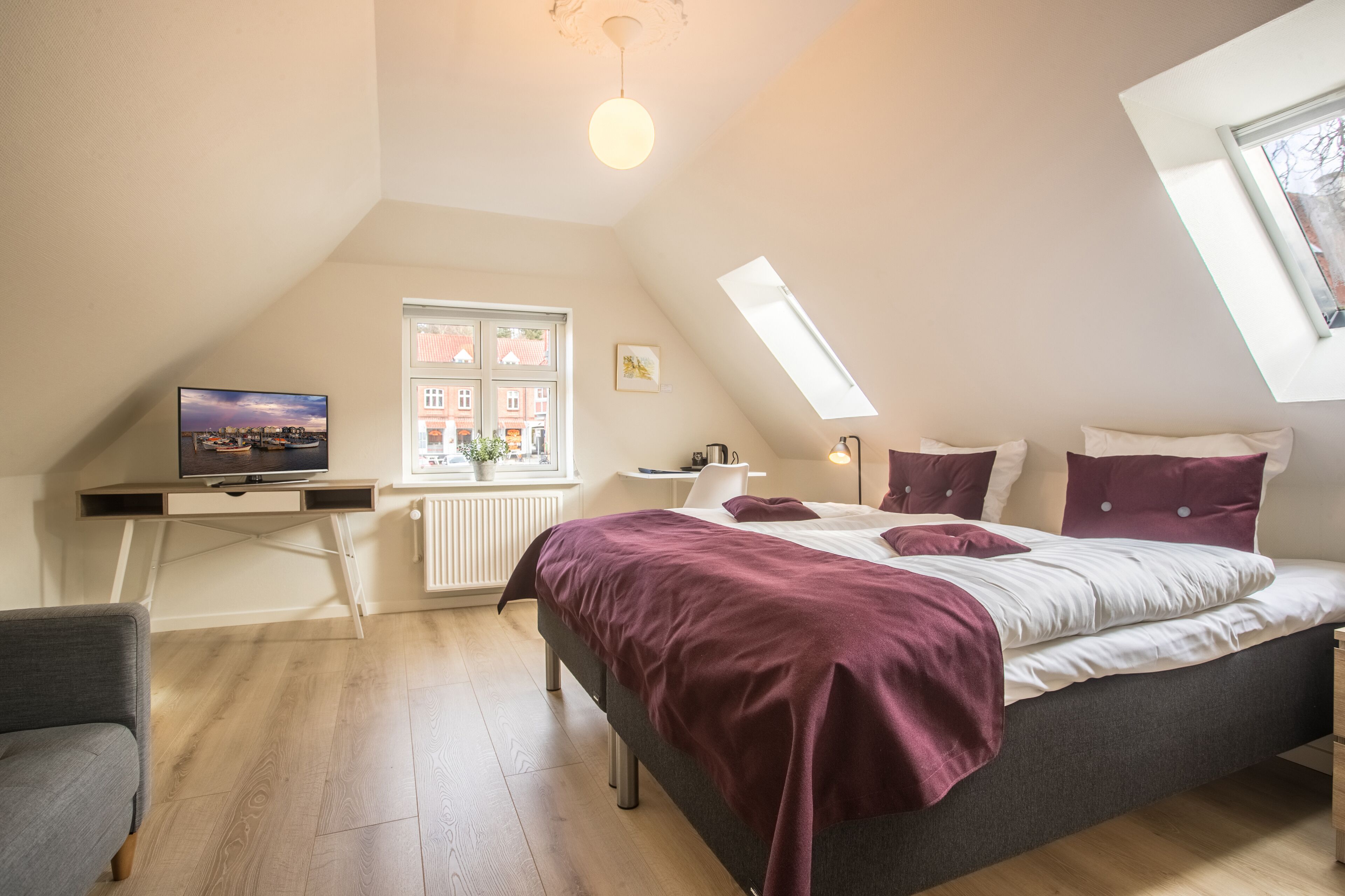 Quarto com cama, escrivaninha, cadeira, televisão e janela com vista para edifícios.