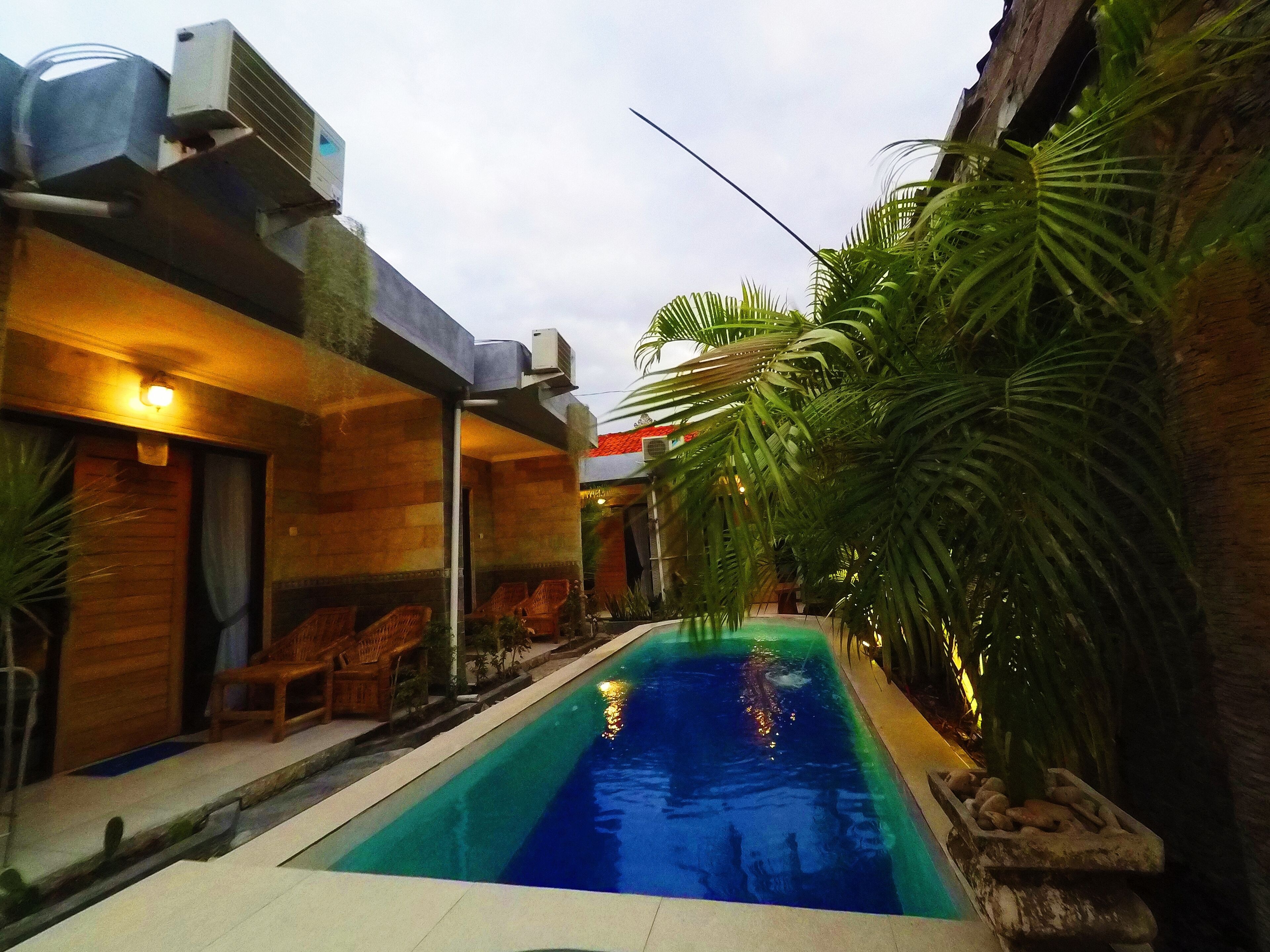 A swimming pool with a wooden deck, surrounded by lush greenery and a modern building with outdoor seating.