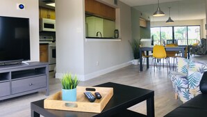 A modern living room with a flat-screen TV, a coffee table with remote controls, a dining area with yellow chairs, and a kitchen with a microwave. - Sunset beach getaway (Treasure Island)