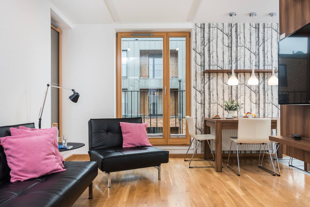 A modern living room with a black leather sofa, a dining table with chairs, and a large window with a view of the city.