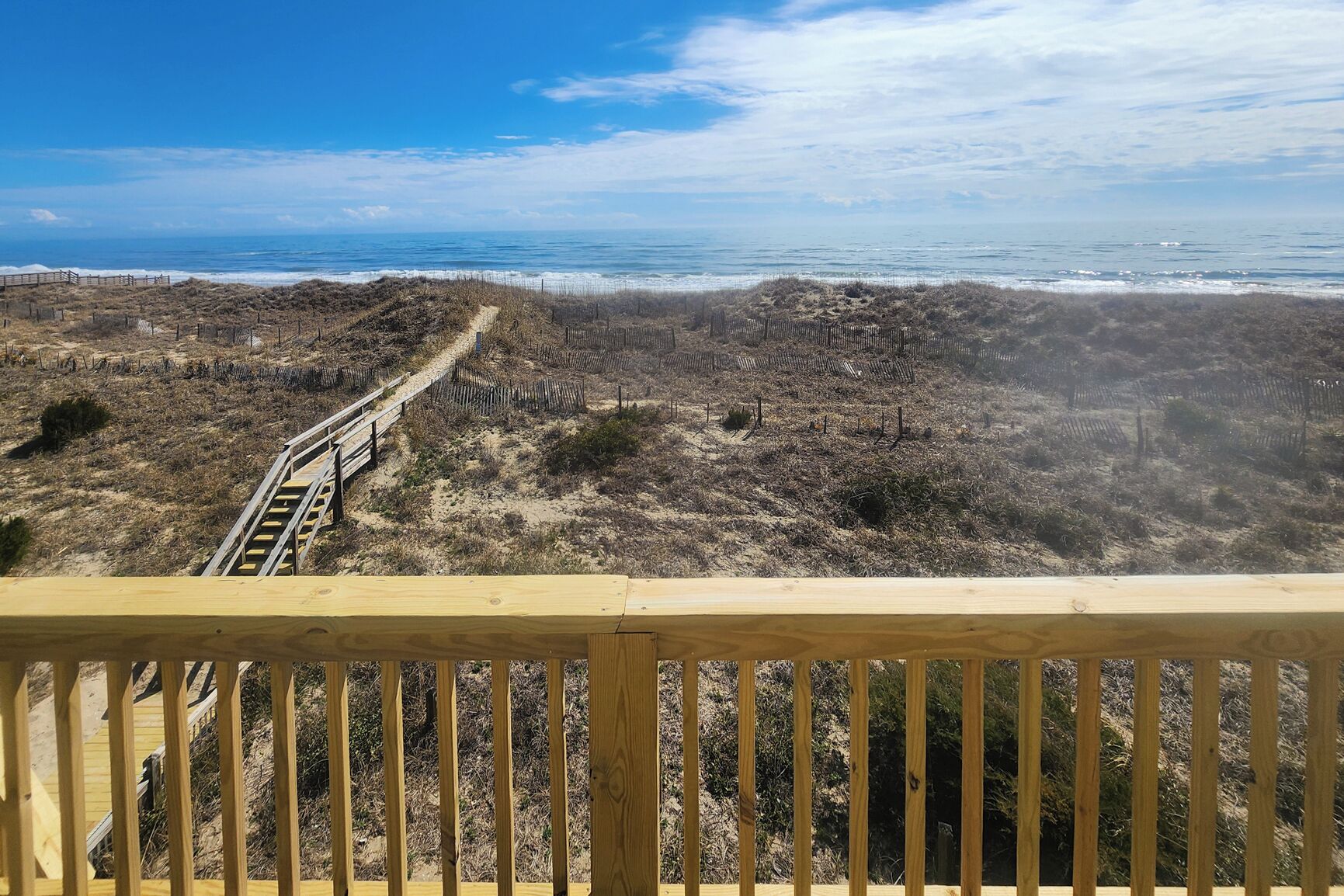 Una terraza de madera con vista a una playa y un sendero arenoso que lleva al océano.