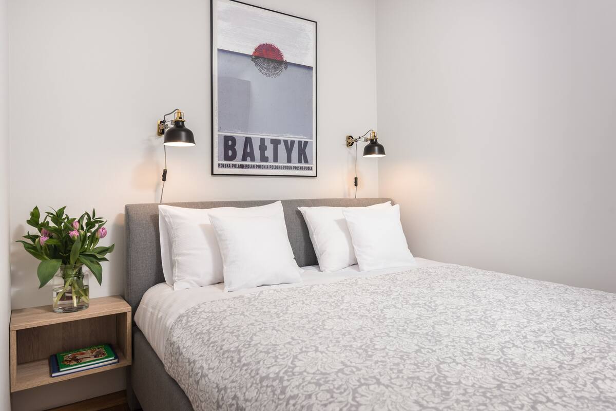 A neatly made bed with a patterned bedspread, a framed poster on the wall, two bedside lamps, a small bedside table with a book and a vase of flowers.