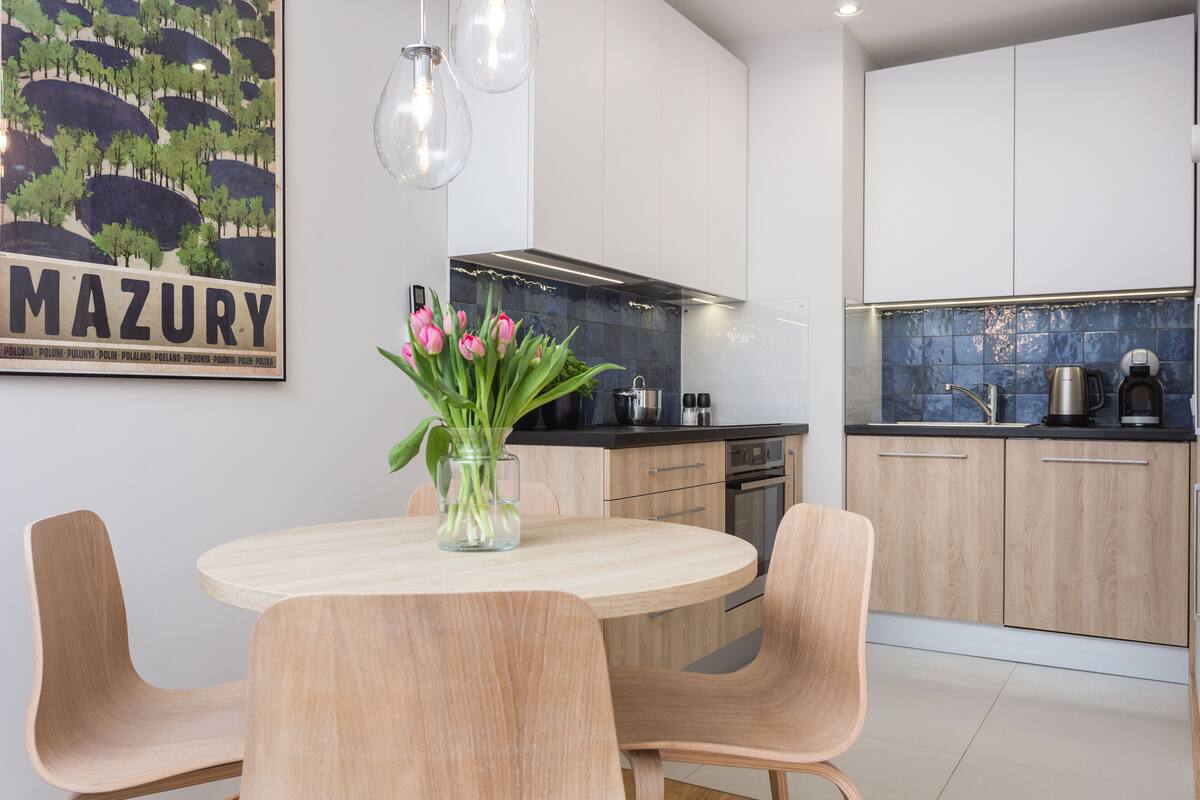 A modern kitchen with light wood cabinets, a black countertop, and a vase with pink tulips on a round table.