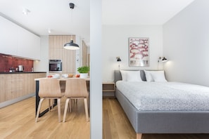 A modern kitchen with wooden cabinets, a dining table with chairs, and a pendant light. There is a bed with a gray bedspread and a framed picture on the wall.