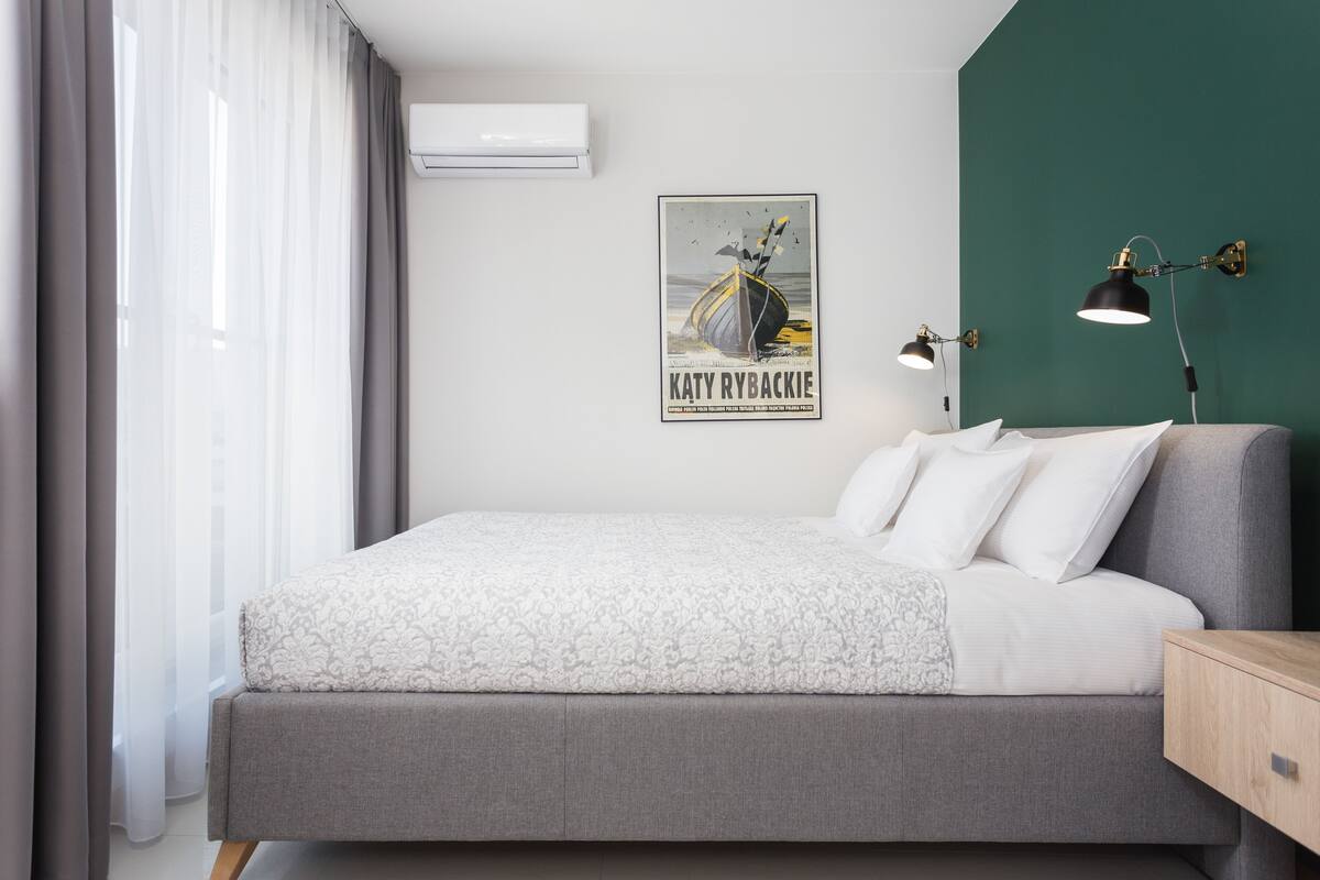 A modern bedroom with a large bed, a wall-mounted air conditioner, a framed poster, and two wall-mounted reading lamps.