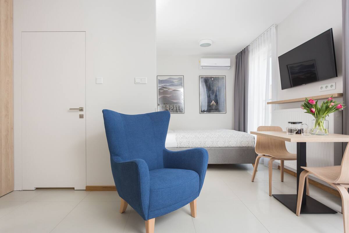 A modern hotel room with a blue armchair, a dining table with chairs, a flat-screen TV, and artwork on the walls.