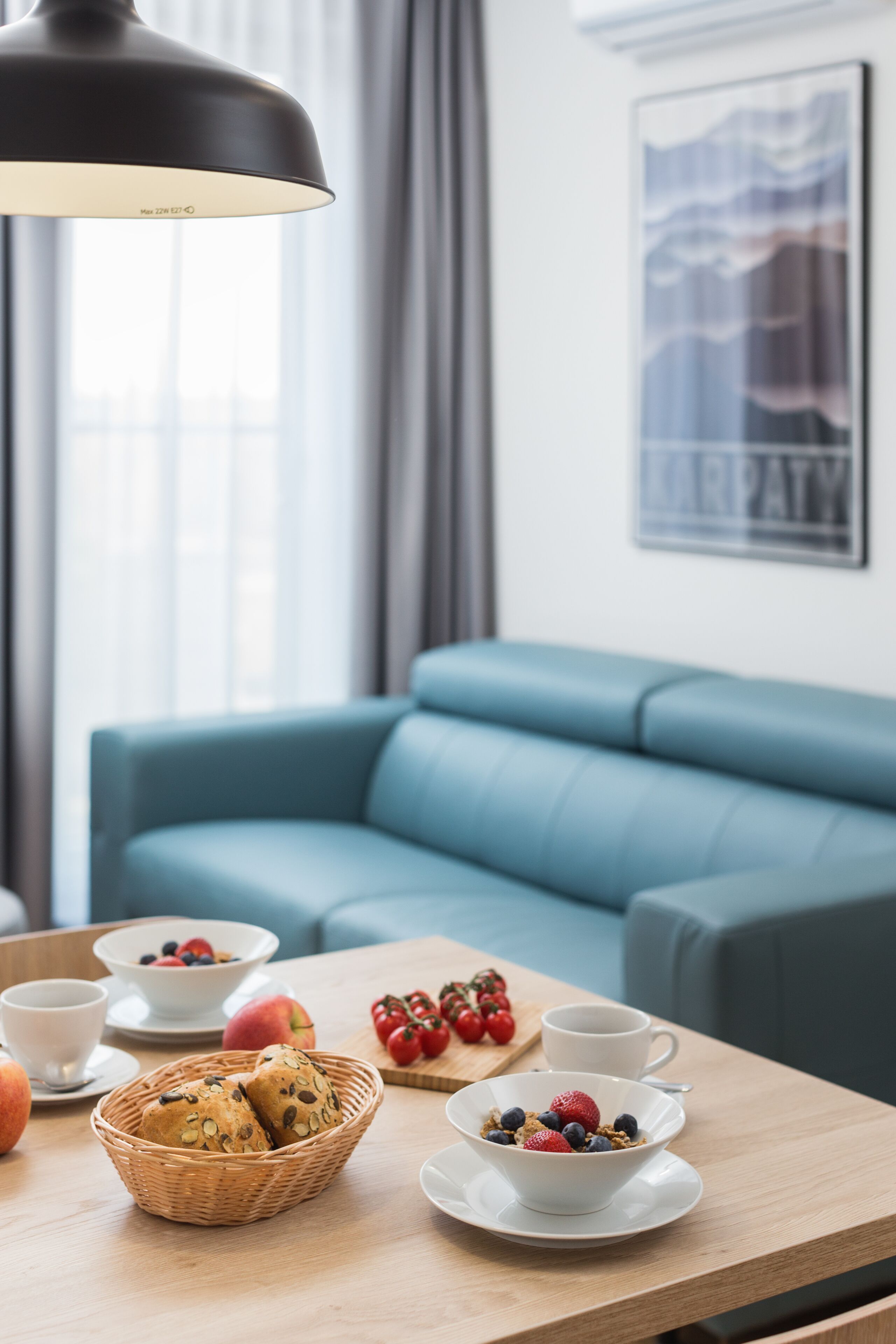 Uma sala de estar moderna com um sofá azul, uma mesa de madeira com itens de café da manhã e uma luminária pendente preta.