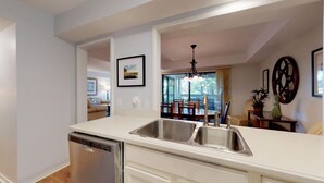 A modern kitchen with white countertops, stainless steel sinks, and a dishwasher. There is a dining area with a table and chairs visible through an open doorway. - Fantastic Fazio Villa with Golf Course View! (Hilton Head)