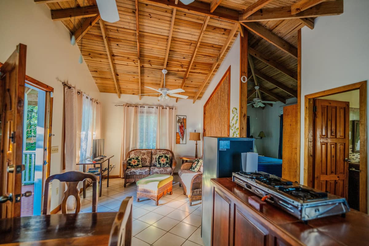 A spacious living area with a high ceiling, wooden beams, a sofa set, a dining table, and a kitchen area with a gas stove.