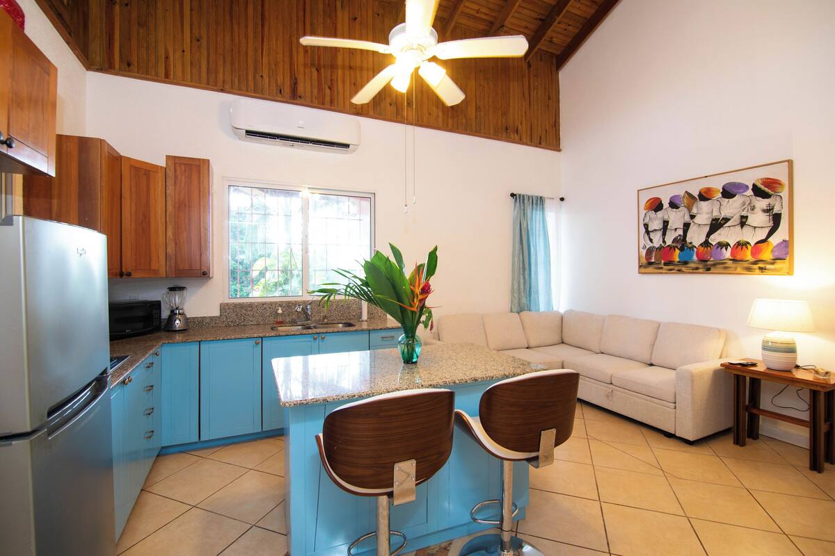 A kitchen with blue cabinets, a refrigerator, a coffee maker, and a dining area with two bar stools.